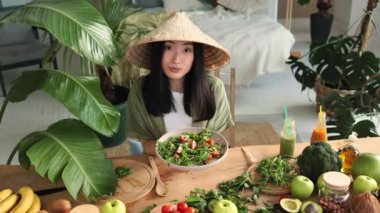 Güzel, genç ve mutlu Asyalı kadın taze organik sebzelerle sağlıklı salata yiyor, yeşil malzemelerle birlikte şık egzotik stüdyoda oturuyor..