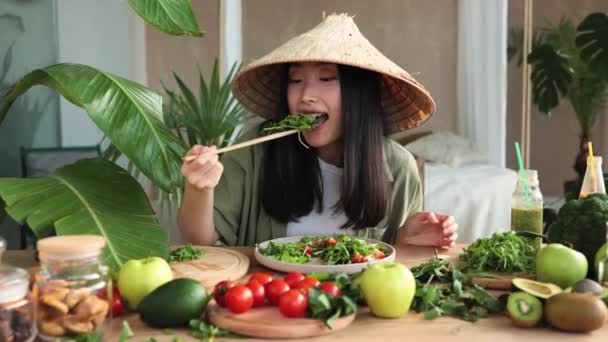 伝統的な円錐形の帽子の陽気でかわいい幸せな若いアジアの女性は 熱帯エキゾチックなリゾートのテーブルの上に座って健康的なサラダを食べています 緑の健康的なビーガンフードコンセプト — ストック動画
