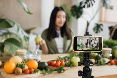Gülümseyen Asyalı genç kadın vlogger pişirirken video blogunu takipçilerine canlı yayınlarken, meyve püresi, elma salatası ve diğer malzemelerin ahşap masada nasıl hazırlandığını gösteriyor.