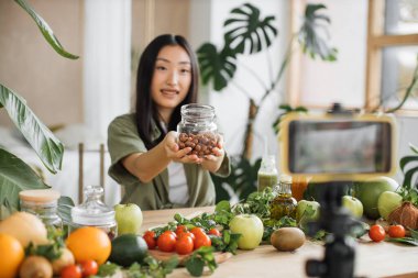 Genç Asyalı kadın blogcu ya da içerik yaratıcısı telefonda salata hazırlıyor ve video kaydediyor. Egzotik mutfakta telefonla blog kaydederken elinde fındık tutan çekici bir kadın.