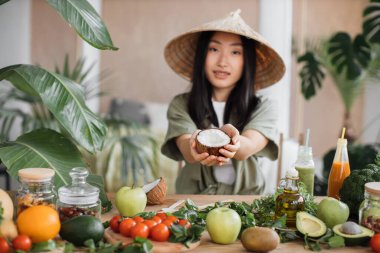 Geleneksel koni şapkalı güzel Asyalı kadın elinde hindistan cevizi, elinde meyve ve sebze malzemeleriyle birlikte şık tropikal mutfakta, kapalı bir masada, sağlıklı vegan salatası yapmak için masada oturuyor..