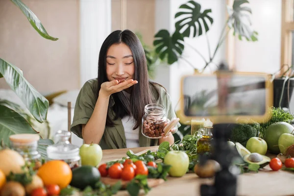 Exotic tropical healthy food concept. Beautiful smiling asian young woman blogger in traditional conical hat cooking fresh organic healthy food using hazelnuts indoor at exotic tropical home studio.