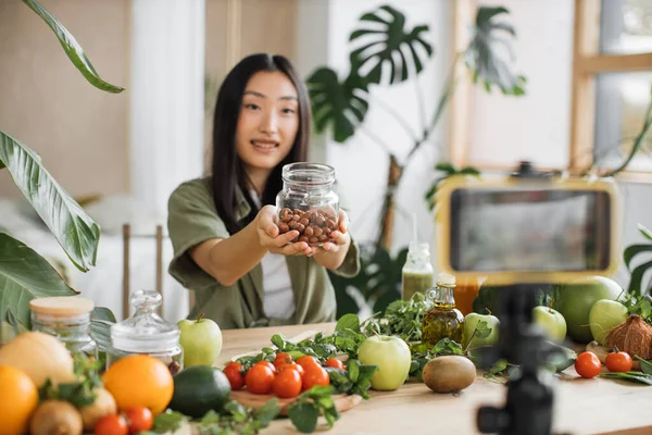 若いアジアの女性ブロガーやコンテンツ作成者は 健康的なサラダを準備し 携帯電話でビデオを記録します 魅力的な女性保持ヘーゼルナッツとともにブログで電話でエキゾチックなキッチン — ストック写真