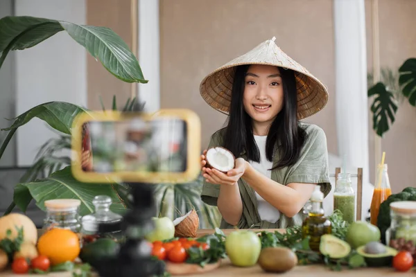 Social media and content creator occupation new job in future. Beauty asian blogger in traditional conical hat using coconut to make smoothies for breakfast and recording video for online channel live