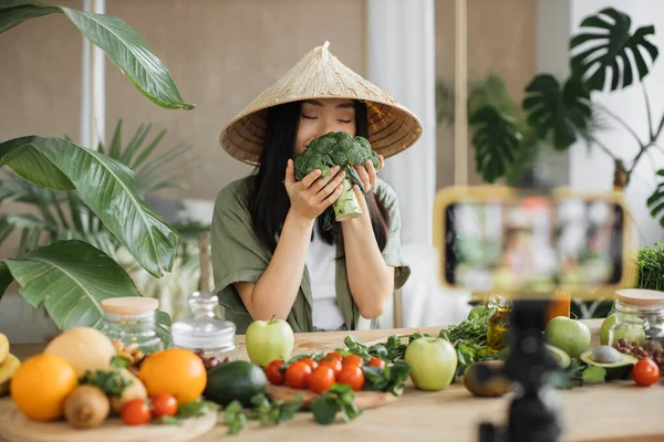Exotic tropical healthy food concept. Beautiful smiling asian young woman blogger in traditional conical hat cooking fresh organic healthy food holding broccoli indoor at exotic tropical home studio.