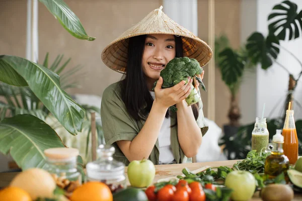 stock image Exotic tropical healthy food concept. Beautiful smiling asian young woman blogger in traditional conical hat cooking fresh organic healthy food holding broccoli indoor at exotic tropical home studio.