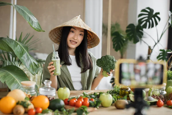 伝統的な円錐形の帽子のアジアの女性料理Vloggerは フォロワーに彼女のビデオブログをライブストリーミングしながら 熱帯リゾートでブロッコリーや他の屋内成分からスムージーを準備する方法を示します — ストック写真