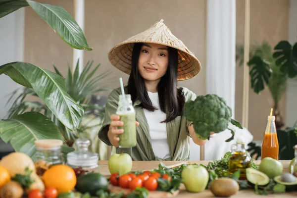 伝統的な円錐形の帽子の楽しいアジアの女性は 熱帯リゾートのエキゾチックな光スタジオで健康的なディナーを楽しんでブロッコリーや他の有機野菜や果物を使用してスムージーを作っています 重量損失 — ストック写真