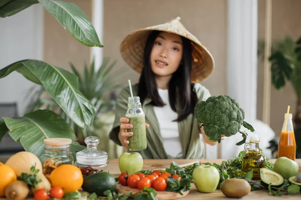 伝統的な円錐形の帽子の楽しいアジアの女性は 熱帯リゾートのエキゾチックな光スタジオで健康的なディナーを楽しんでブロッコリーや他の有機野菜や果物を使用してスムージーを作っています 重量損失 — ストック写真