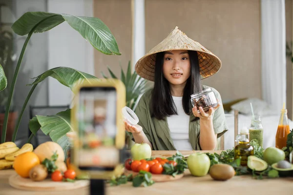 Exotic tropical healthy food concept. Beautiful smiling asian young woman blogger in traditional conical hat cooking fresh organic healthy food using dates indoor at exotic tropical home studio.