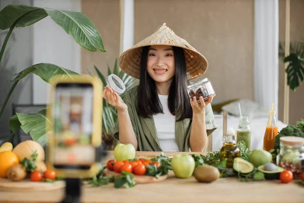 Exotic tropical healthy food concept. Beautiful smiling asian young woman blogger in traditional conical hat cooking fresh organic healthy food using dates indoor at exotic tropical home studio.