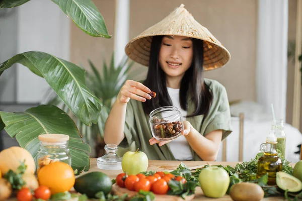 免疫力を高めるための健康食品 美しい笑顔アジアの若い女性ブロガーで伝統的な円錐形の帽子調理新鮮な有機健康食品使用して日付屋内でエキゾチックなトロピカルホームスタジオ — ストック写真