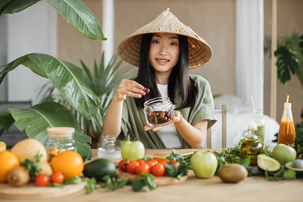 免疫力を高めるための健康食品 美しい笑顔アジアの若い女性ブロガーで伝統的な円錐形の帽子調理新鮮な有機健康食品使用して日付屋内でエキゾチックなトロピカルホームスタジオ — ストック写真