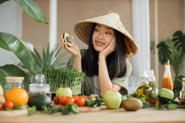 トロピカルリゾートのエキゾチックな光スタジオでアボカドと健康的なディナーを楽しむ野菜や果物のサラダを作る伝統的な円錐形の帽子で楽しいアジアの女性 減量のための食品や栄養 — ストック写真