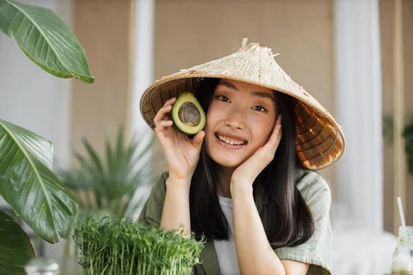 健康的なビーガンサラダを作るために果物や野菜成分とテーブルに座ってアボカドを保持伝統的な円錐形の帽子の美しいアジアの女性 スタイリッシュなトロピカルキッチンでテーブルの屋内 — ストック写真