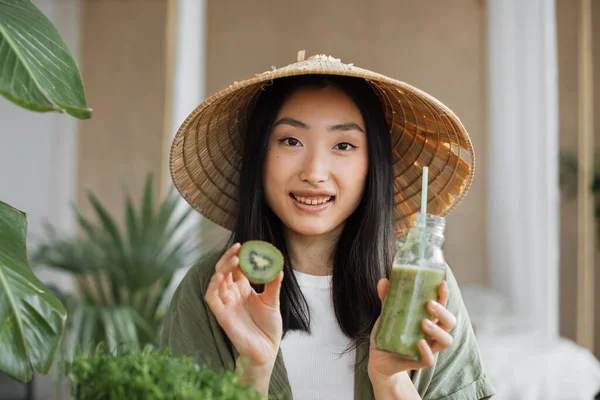 stock image Young asian woman in conical hat holding in her hands glass bottle with healthy tonic smoothie and kiwi while sitting on table with green fresh ingredients indoors in stylish light tropical studio.