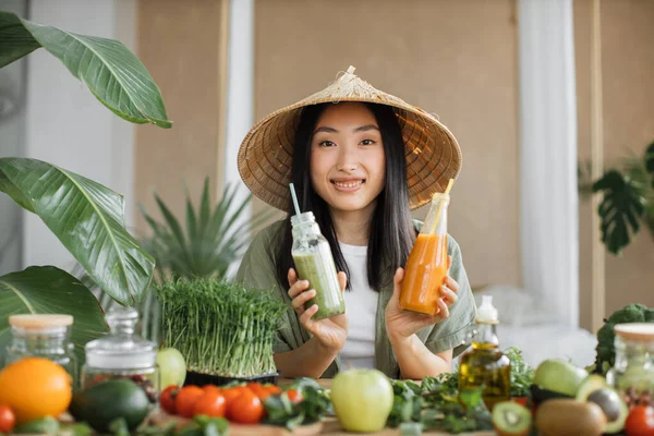 Charming young asian vegan woman preparing healthy fresh smoothie for breakfast on tropical exotic kitchen. Attractive female holding in hands two bottle with smoothies. Green healthy food concept.