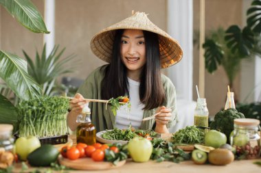 Geleneksel konik şapkalı neşeli mutlu Asyalı kadın tropik egzotik tatil beldesinin kapalı masasında oturmuş sağlıklı salata yiyor. Yeşil sağlıklı vegan besin konsepti.