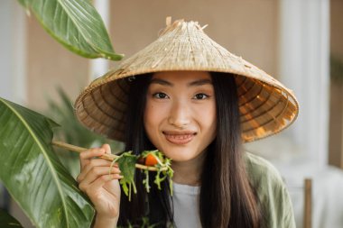 Güzel, genç ve mutlu Asyalı kadın taze organik sebzelerle sağlıklı salata yiyor, yeşil malzemelerle birlikte şık egzotik stüdyoda oturuyor..
