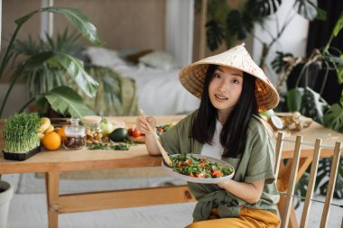 Geleneksel konik şapka takan ve sağlıklı salata yiyen güzel, mutlu Asyalı bir kadın. Masanın yanında, organik sebzeler ve meyve malzemeleriyle birlikte, tropikal tatil köyünde oturuyor..