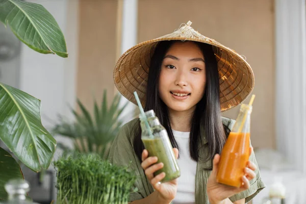 Charming young asian vegan woman preparing healthy fresh smoothie for breakfast on tropical exotic kitchen. Attractive female holding in hands two bottle with smoothies. Green healthy food concept.