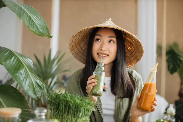 伝統的な円錐形の帽子の若いアジアの女性は スタイリッシュな光のトロピカルスタジオで室内で緑の新鮮な食材とテーブルの上に座っている間 わらでボトルから健康的な強壮スムージーを飲む — ストック写真