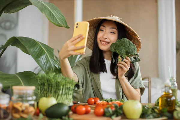 Social media and content creator occupation new job in future. Beauty asian blogger in traditional conical hat holding phone and broccoli to make healthy food for breakfast