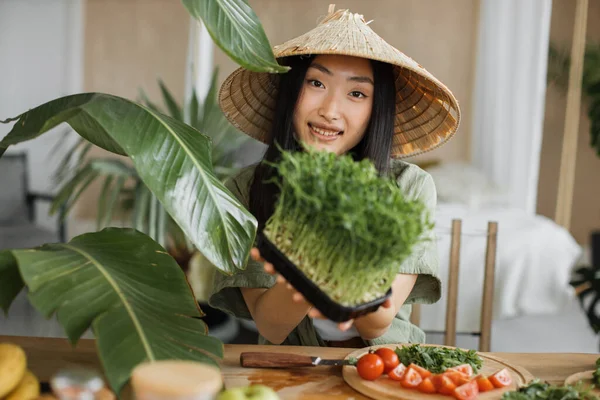 若い魅力的なアジアの女性伝統的な円錐形の帽子でテーブルに座ってスタイリッシュな光トロピカルキッチン健康的な野菜サラダを作るためにマイクログリーンの芽を保持 — ストック写真