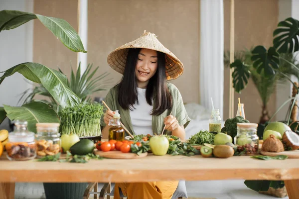 伝統的な円錐形の帽子の美しいアジアの女性は 熱帯エキゾチックなキッチンで木製のスプーンを使用して新鮮な野菜と新鮮な健康的なビーガンサラダを攪拌 自宅でスタジオ 新しいレシピを試してみてください — ストック写真