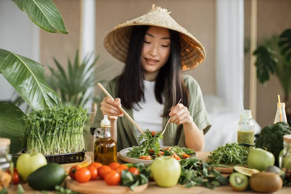 伝統的な円錐形の帽子の美しいアジアの女性は 熱帯エキゾチックなキッチンで木製のスプーンを使用して新鮮な野菜と新鮮な健康的なビーガンサラダを攪拌 自宅でスタジオ 新しいレシピを試してみてください — ストック写真