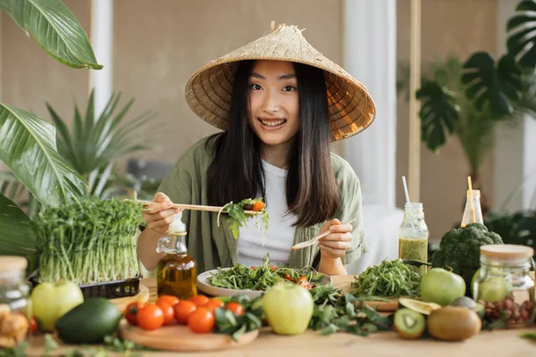 伝統的な円錐形の帽子の陽気でかわいい幸せな若いアジアの女性は 熱帯エキゾチックなリゾートのテーブルの上に座って健康的なサラダを食べています 緑の健康的なビーガンフードコンセプト — ストック写真