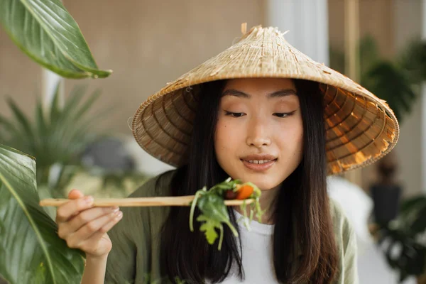 stock image Beautiful young and happy asian woman eating healthy salad with fresh organic vegetables, sitting on table with green fresh ingredients at stylish light exotic studio.