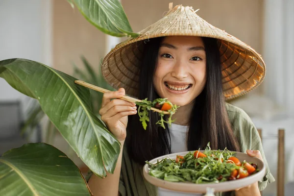 美しく若いと幸せなアジアの女性は スタイリッシュな光エキゾチックなスタジオで緑の新鮮な食材とテーブルの上に座って 新鮮な有機野菜と健康的なサラダを食べる — ストック写真