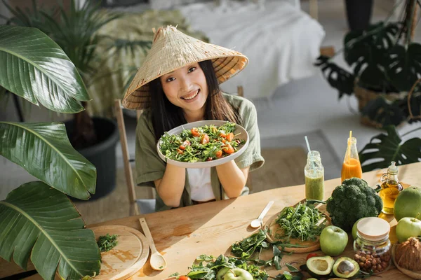 伝統的な円錐形の帽子の中で楽しいアジアの女性は 熱帯リゾートのエキゾチックな光スタジオで健康的なディナーを楽しんでいる有機野菜や果物からサラダを保持しています 減量のための食品や栄養 — ストック写真
