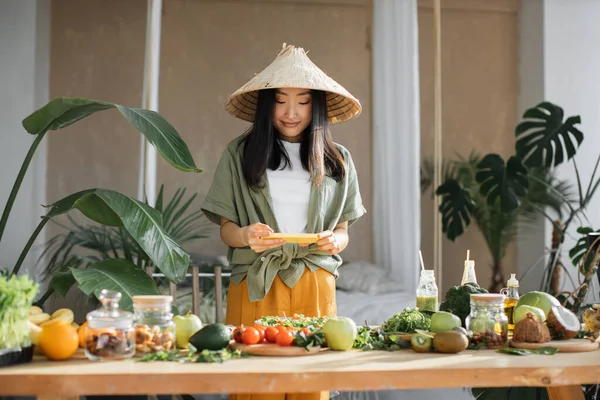 若いアジアのブロガーの女性は ソーシャルメディアに写真を投稿するためにスマートフォンを使用して キッチンに立って エキゾチックな家でサラダを作る女性の伝統的な円錐形の帽子 家庭的なコンセプトでリラックスしたライフスタイルの女性 — ストック写真