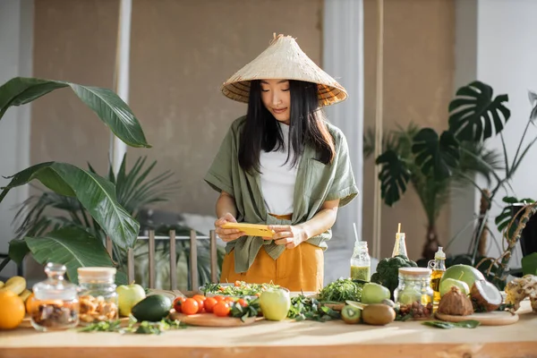 Young asian blogger woman using smartphone to posting photos in social media, standing in kitchen, female traditional conical hat making salad at exotic home. Lifestyle women relaxing at home concept.