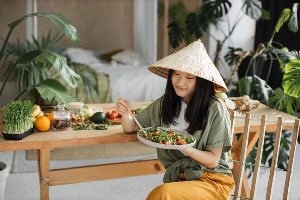 伝統的な円錐形の帽子の美しい幸せな若いアジアの女性有機野菜や果物の成分を屋内熱帯リゾートでテーブルの近くに座って健康的なサラダを保持し 食べる — ストック写真