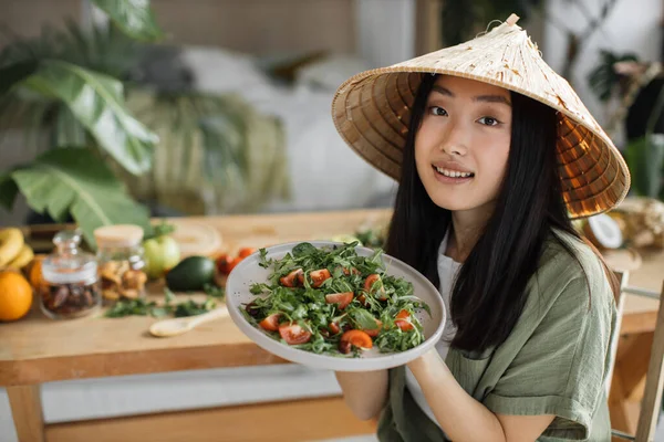 トロピカルリゾートのエキゾチックな光スタジオで健康的なディナーを楽しむ有機野菜から健康的なサラダを保持している伝統的な円錐形の帽子の楽しいアジアの女性 減量のための食品や栄養 — ストック写真