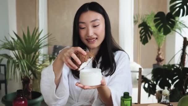 ココナッツの半分を保持し 健康的な肌のための自家製クリームを作る若いアジアの女性 木製のテーブルの上に横たわる自家製化粧品のための多くの成分 — ストック動画