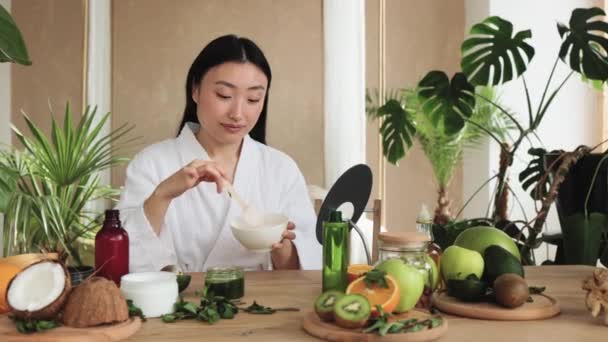 白いバスローブを着た美しい健康的なアジアの女性は クリームを準備し 顔に適用しながら ボウルに天然化粧品を調製様々な成分と木製のテーブルに座っています — ストック動画