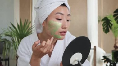 Masada elinde ev yapımı kozmetik malzemeleriyle oturan çekici Asyalı kadın yüzüne yeşil nemlendirici maske takıyor. Genç bayan yaşlanma karşıtı prosedürler uyguluyor..