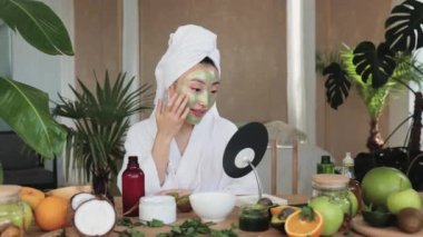 Yeşil nemlendirici kozmetik maskeli ve tropik arka planda aynaya bakan Asyalı genç bir kadının portresi. Cilt bakımı için düzenli güzellik tedavisi.