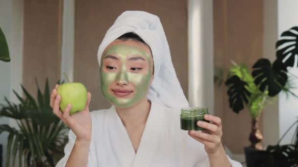 緑のフェイスマスクをした頭の上にバスタオルを持つ快適なアジアの女性は トロピカルビューの背景を持つ緑の物質室内でリンゴとガラス製品を保持しています スキンケアコンセプト — ストック動画