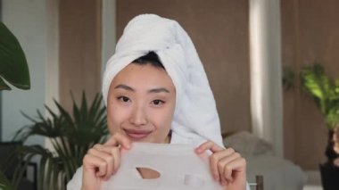 Beyaz havluyla kameraya gülümseyen, elinde pamuk maskeyle tropikal egzotik stüdyo arkaplanı olan Asyalı güzel bir kadının portresi. Yüz bakımı ve güzellik anlayışı.