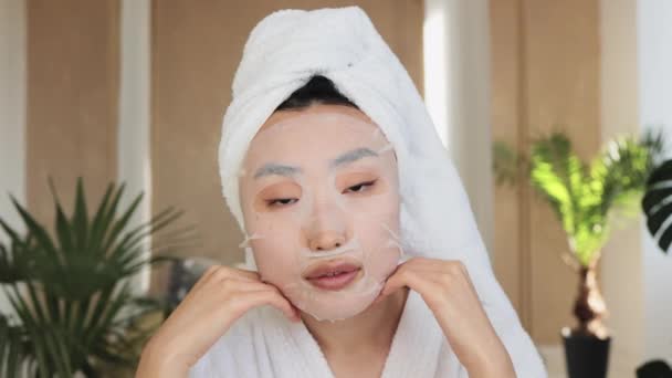 エキゾチックな背景の上に頭の上に綿マスクシートとタオルと美しい若いアジアの女性の肖像画 かなり女性行うスキンケア手順室内で自宅 — ストック動画