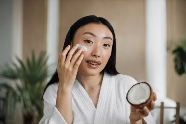 Genç Asyalı kadının evde doğal kozmetik uyguladığı, cilt için ev yapımı krem, kaplıca merkezinde egzotik bir geçmişi olan saç bakımı için hindistancevizi tutan yüzünün yakın çekimi.