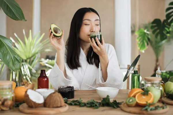 在用新鲜鳄梨制作天然手工化妆品的混合物时 快乐的亚洲女人拿着融化的绿色肥皂团玻璃器皿 或在桌上涂上保湿滋养面膜 — 图库照片