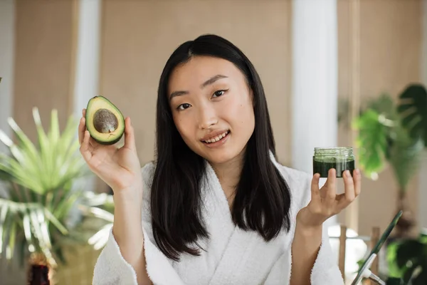 Blogger or content creator asian woman preparing natural cosmetics at home holding avocado homemade cream in glassware for skin and hair care home spa sitting at wooden table with exotic background