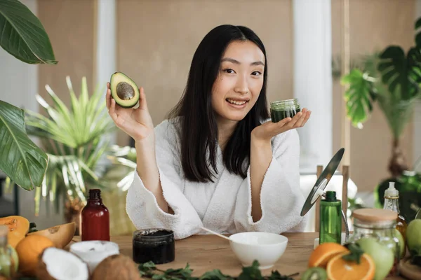 アボカドを持っている若いアジアの女性と健康的な肌のための自家製クリームを作る 木製のテーブルの上に横たわる自家製化粧品のための多くの成分 — ストック写真