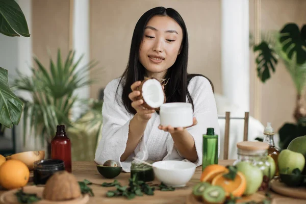 stock image Focus on coconut and glass jar, young asian woman holding half of coconut and making homemade cream